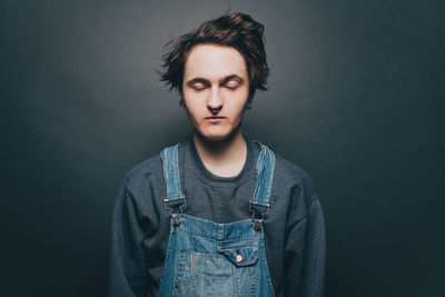 Young man with eyes closed wearing denim overalls over gray background
