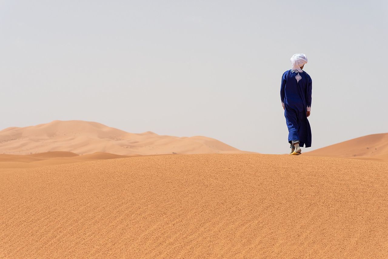 REAR VIEW OF MAN STANDING ON SAND DUNE