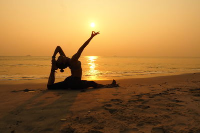 Full length of woman practicing yoga on shore at beach against sky during sunset