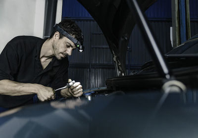 Technician with head flashlight using ratchet while repairing motor of automobile in car station