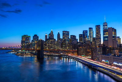 Illuminated bridge over river by buildings against blue sky