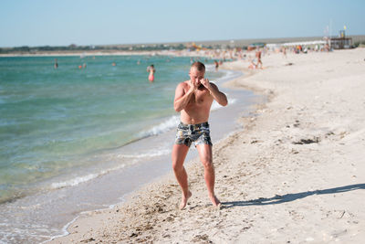 Full length of shirtless man boxing at beach against sky