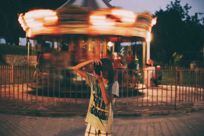Young woman photographing while standing against illuminated carousel during sunset