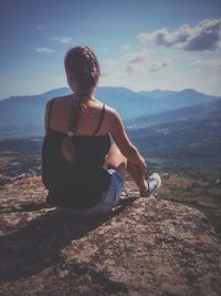 Rear view of woman sitting on cliff against sky during sunny day