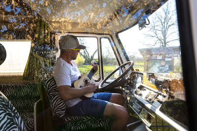 Side view of man playing guitar while sitting in bus seen through window