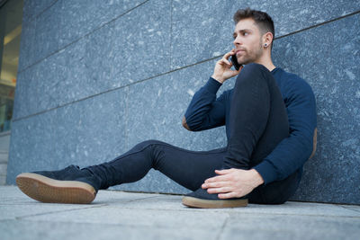 Side view of modern male in casual wear sitting near stone wall and br