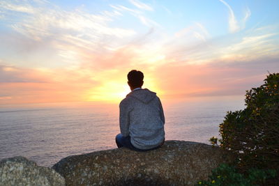 Rear view of man sitting on rock by sea against sky during sunset