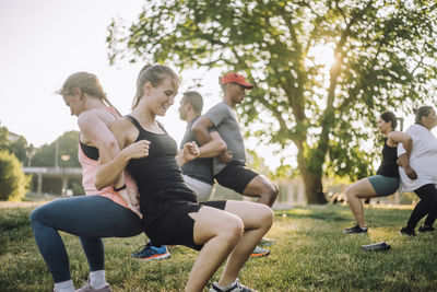 Smiling multiracial male and female friends doing buddy back stretch on grass at park