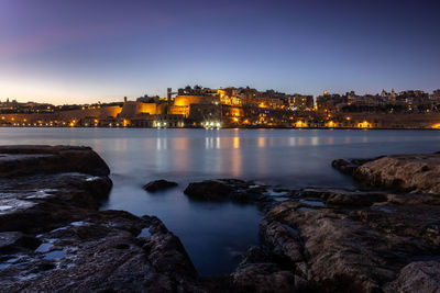 Illuminated buildings by sea against sky at dusk