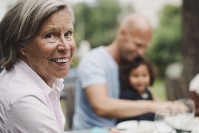 Side view portrait of happy senior woman with family having food at outdoor table in background