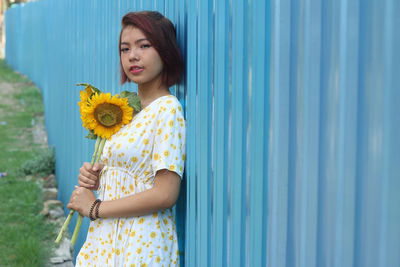 Portrait of woman holding sunflowers while leaning on corrugated iron