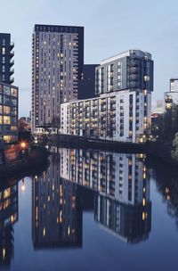 Illuminated buildings by river against sky in city at dusk