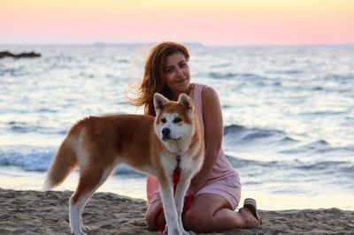 Portrait of smiling young woman with dog at beach against sky during sunset