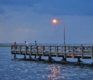 People fishing while standing on pier during sunset