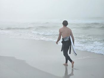 Surfer from behind walking down a foggy beach.