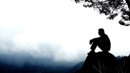 Silhouette man sitting on rock against sky