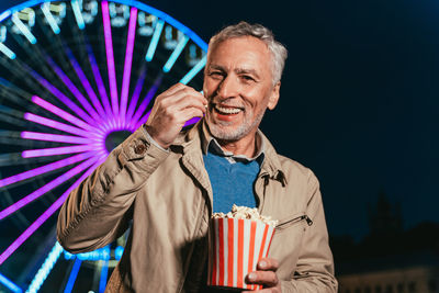 Portrait of smiling man eating popcorn standing against ferris wheel