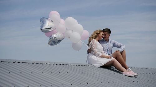 Young couple with balloons sitting on roof against sky