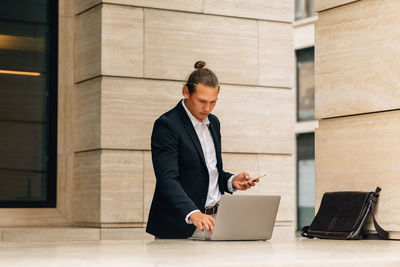 Businessman using laptop while holding smart phone against building