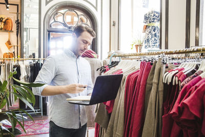 Confident male entrepreneur using laptop while standing by clothes rack in boutique