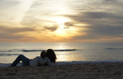 Couple relaxing at beach against sky during sunset