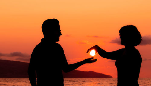 Couple sitting at beach, silhouette in sunset, love and friendship, woman holding the sun.