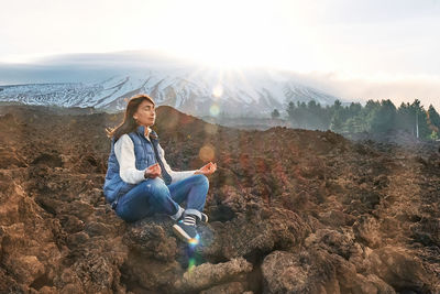 Woman enjoying freedom while meditating on lava stone at panoramic view of snowy summits of etna