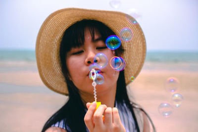 Close-up of young woman wearing hat blowing bubbles while standing against sky