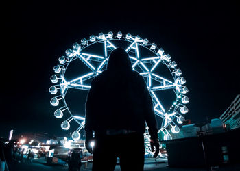 Rear view of silhouette man standing against illuminated lights at night