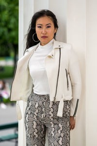 Young mongolian woman with a white leather jacket and patterned well-fitting pants in an urban park.