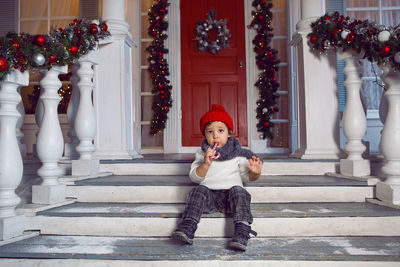 Baby boy sitting on the steps of the house with snow and eating striped candy christmas