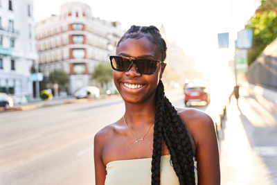 Happy african american female in stylish clothes and sunglasses with long braids smiling and looking at camera while standing near road on city street