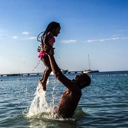 Side view of smiling father lifting daughter while standing in sea against sky