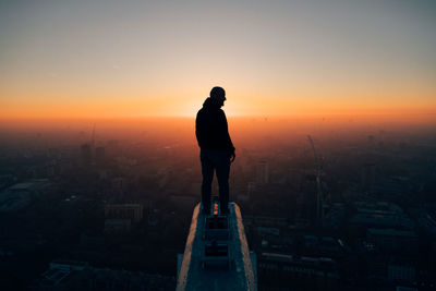 Rear view of silhouette man looking at cityscape against sky during sunset