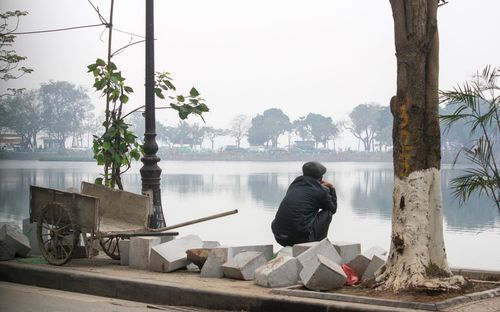 Rear view of man crouching at lakeshore against sky