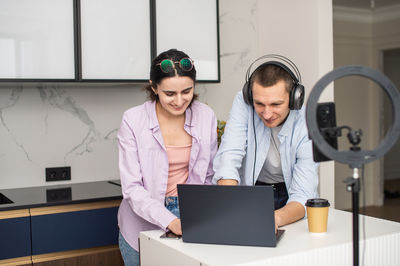 A man with headphones and a girl with glasses are streaming and looking at a laptop in the room