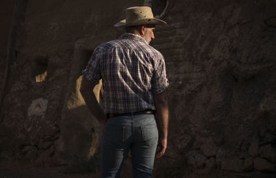 Rear view of adult man in cowboy hat and shirt against abandoned building