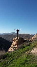 Rear view of young man with arms outstretched standing on mountain against clear blue sky