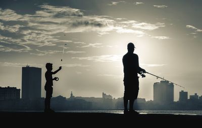 Silhouette father and son fishing in river against sky during sunset at city