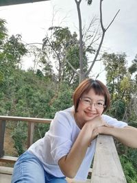 Portrait of smiling young woman sitting on railing against trees