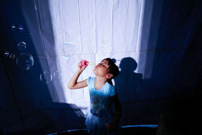 High angle view of girl blowing bubbles by white curtain