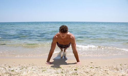 Portrait of shirtless man doing push-ups at beach against clear sky