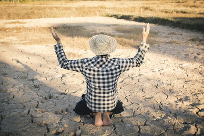 Rear view of person with arms raised kneeling on cracked field