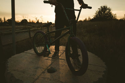 Low section of man with bicycle standing on land during sunset