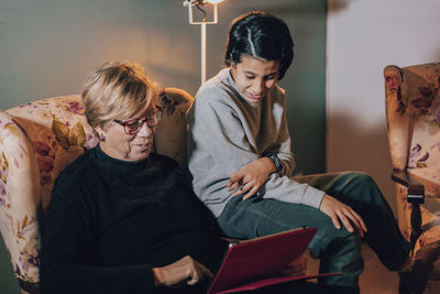 Caucasian grandmother teaches a game on her tablet to her granddaughter sitting on an armchair