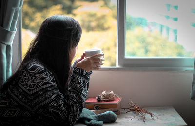 Side view of young woman drinking coffee while looking through window at home