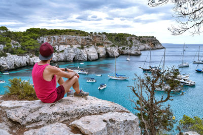 Rear view of man sitting on rock looking at sea against sky