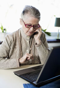 Senior woman using mobile phone while sitting with laptop on table