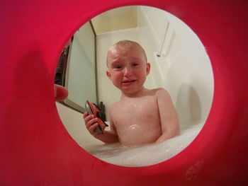 Portrait of shirtless baby boy crying in bathtub seen through red toy