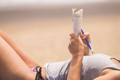 Midsection of woman holding book and pen while lying at beach during sunny day